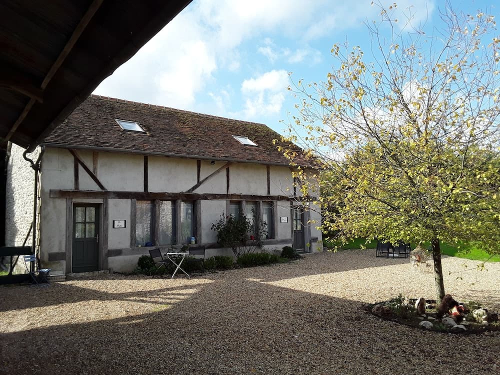 A serene rural retreat features a charming half-timbered cottage with a gravel courtyard, surrounded by lush greenery under a bright blue sky, ideal for a peaceful countryside getaway.
