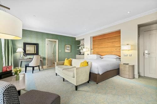 A cozy hotel suite features a plush bed with wooden headboard, a comfortable seating area, and soft green walls, creating a serene atmosphere ideal for relaxation during business or leisure trips.