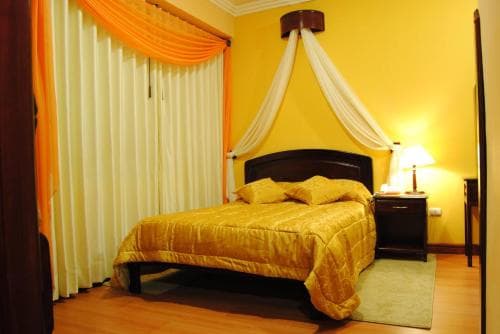 A cozy hotel room features a warmly lit bed with yellow bedding, a canopy draped over the headboard, orange curtains, and a bedside lamp, ideal for travelers seeking comfort and relaxation.
