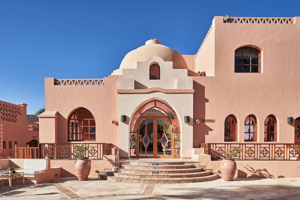A serene courtyard features a charming pink building with arched windows and a dome under a clear blue sky, ideal for travelers seeking a tranquil Middle Eastern retreat experience.