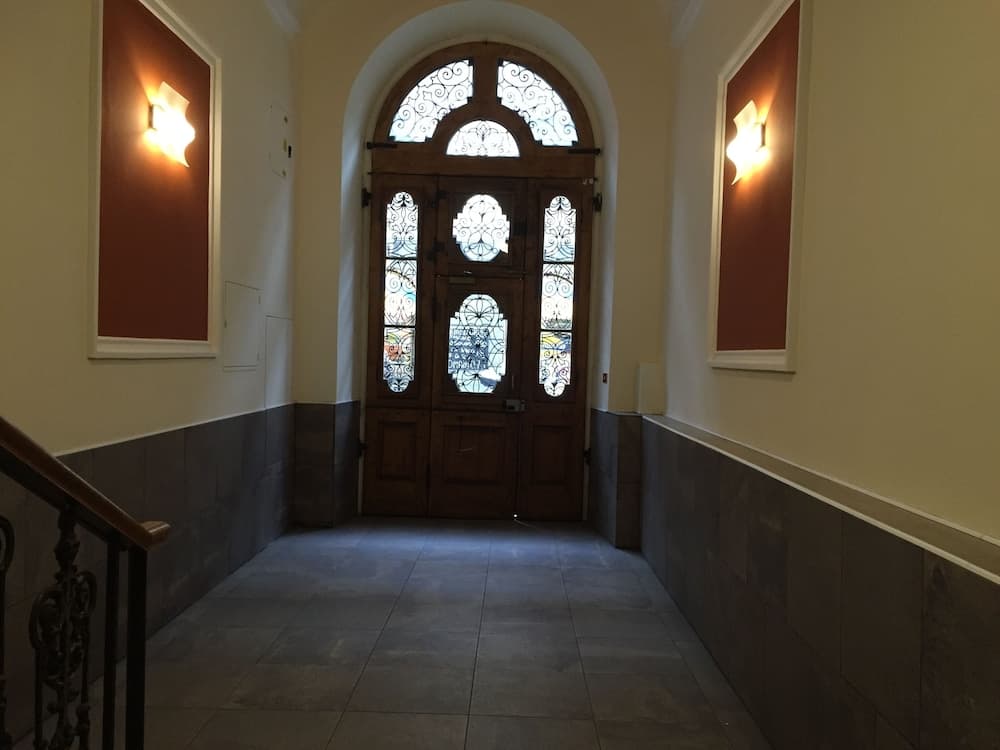 A serene hallway with an ornate arched wooden door featuring stained glass panels, flanked by warm wall sconces and framed artwork, creates an inviting atmosphere ideal for exploring historic buildings or museums.