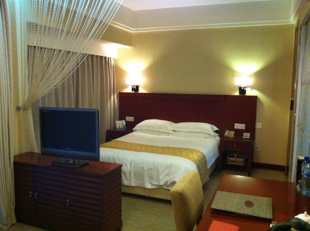 A cozy hotel room features a neatly made bed with white linens, warm lighting from wall-mounted lamps, a flat-screen TV on a wooden dresser, and a serene ambiance ideal for travelers seeking relaxation.