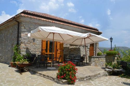 A charming stone villa with a rustic patio shaded by large umbrellas invites travelers to relax amidst vibrant flowers and scenic mountain views, perfect for a serene getaway.