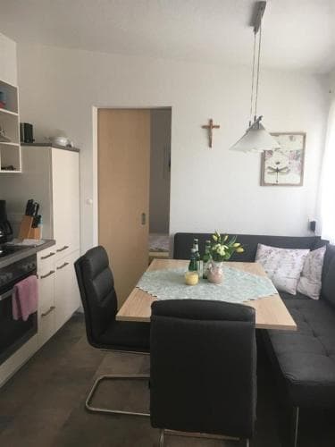 A cozy apartment dining area features a wooden table set with flowers, black chairs, a modern kitchenette, and a comfortable sofa, creating an inviting atmosphere ideal for short-term rentals or family stays.