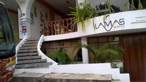 A tropical hotel entrance featuring a curved staircase, lush palm plants, and a sign reading "LAMAS SUITES," ideal for travelers seeking a cozy and inviting accommodation in a warm climate destination.