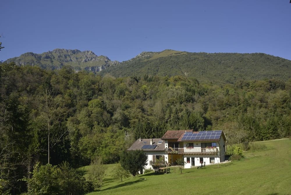 A serene countryside retreat features a charming house with solar panels nestled amidst lush greenery and towering mountains, ideal for eco-conscious travelers seeking tranquility and natural beauty.