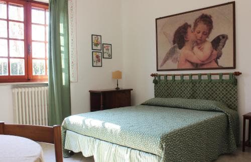 A cozy bedroom with a neatly made bed adorned in green patterns, framed by a serene painting of angels, red-framed windows, and soft lighting, evokes a tranquil European retreat ideal for travelers seeking relaxation.