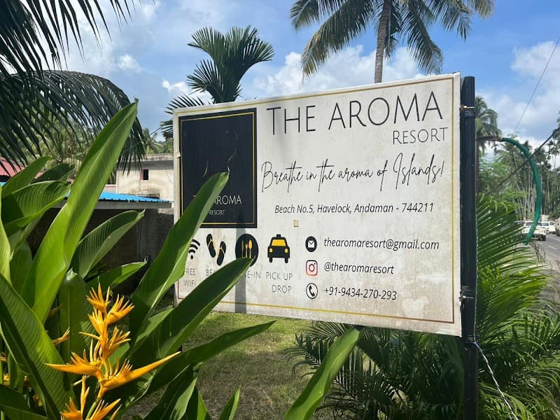 A vibrant signboard for The Aroma Resort amidst lush tropical foliage and palm trees invites travelers to relax on Beach No.5 in Havelock, Andaman and Nicobar Islands, promising an island escape with amenities like Wi-Fi and pickup services.