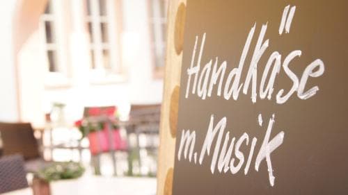 A cozy outdoor café scene features a chalkboard sign reading "Handkäse m. Musik," inviting travelers to enjoy artisanal cheese with music in a relaxed, sunny setting.