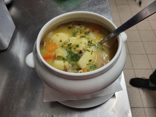 A hearty bowl of chunky potato soup garnished with fresh herbs sits invitingly on a stainless steel countertop in a cozy kitchen setting, perfect for travelers seeking comfort food during their culinary adventures.