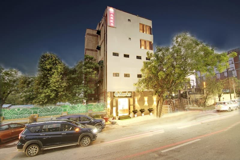 A cozy, well-lit hotel with "Marks" signage stands invitingly on a quiet street at dusk, surrounded by lush greenery and parked cars, ideal for travelers seeking comfort in Noida's bustling industrial area.