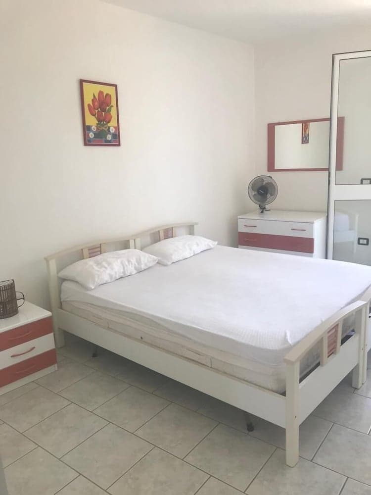 A cozy bedroom with white walls, a neatly made bed, pink-accented furniture, and vibrant artwork creates a serene atmosphere ideal for travelers seeking a comfortable and minimalist accommodation.