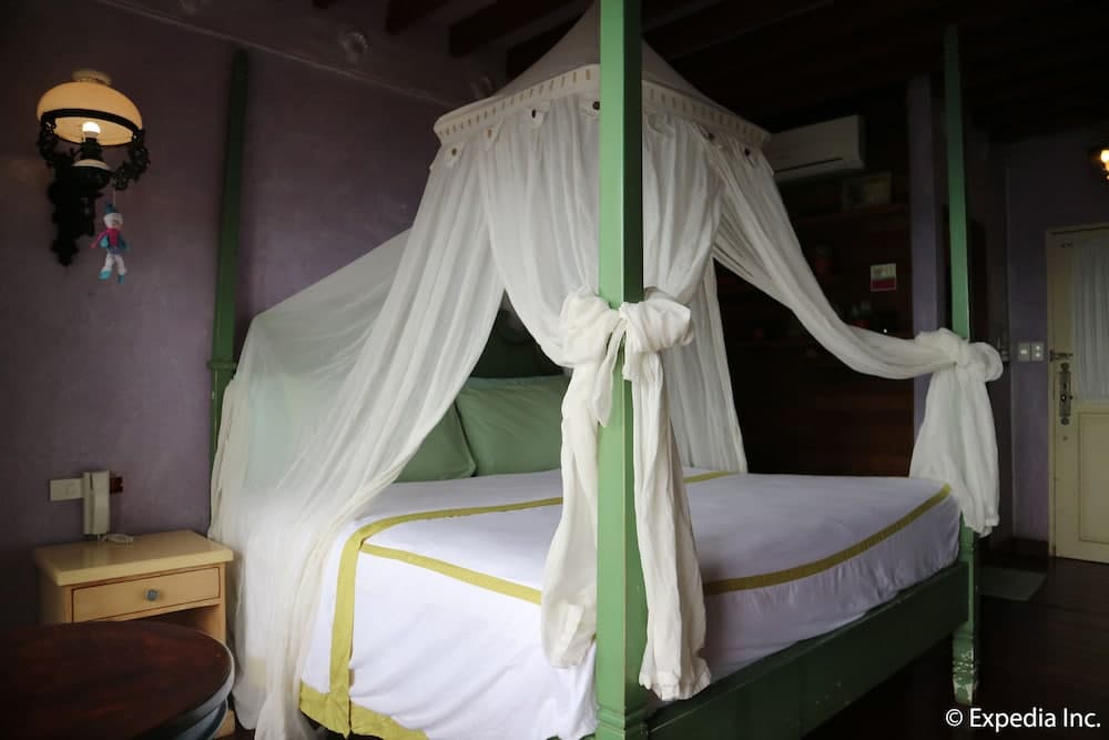 A cozy hotel room features a charming canopy bed draped in white curtains, purple walls, and vintage-style lighting, ideal for travelers seeking a romantic and serene getaway.