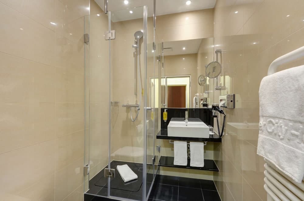 A modern hotel bathroom features beige tiled walls, a sleek glass-enclosed shower, a black vanity with white sink, and neatly arranged towels, ideal for travelers seeking comfort and cleanliness.