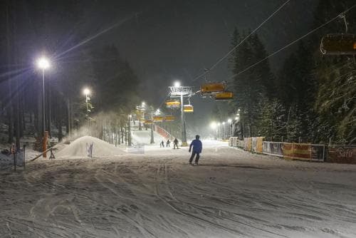 A serene night scene at a ski resort illuminated by bright lights, featuring snow-covered slopes, a chairlift, and scattered skiers, ideal for winter sports enthusiasts seeking a peaceful evening adventure.