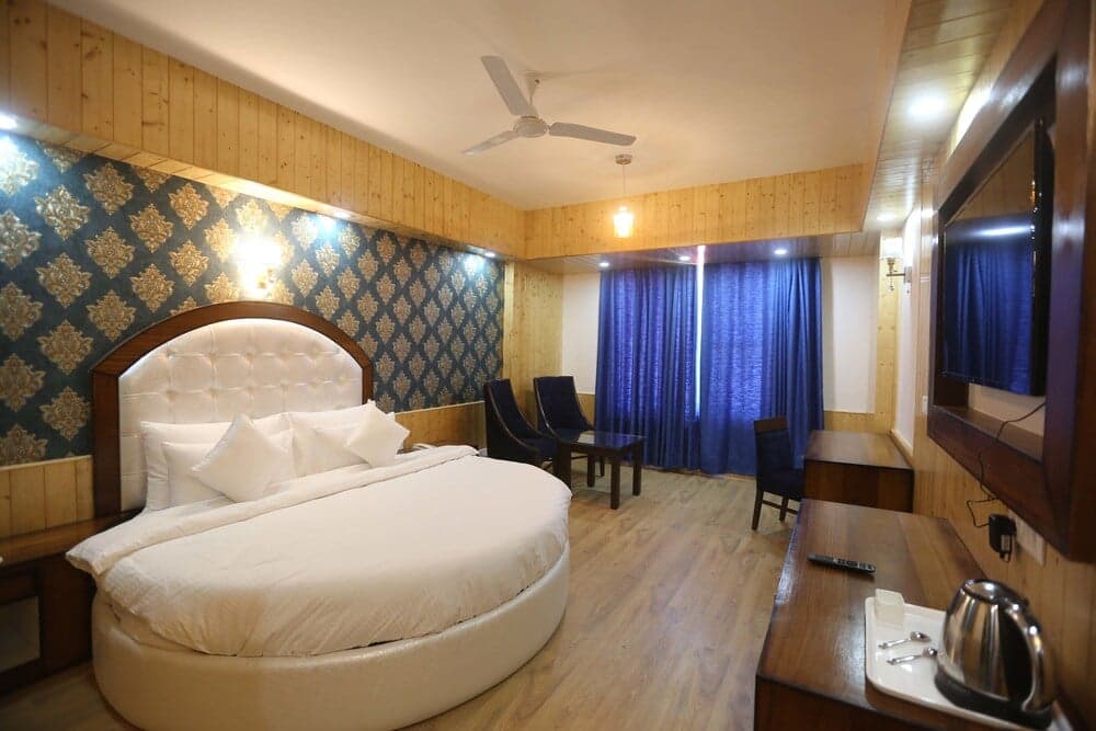 A cozy hotel room features a round bed with white linens, blue curtains, wooden paneling, and modern amenities, creating a warm and inviting atmosphere ideal for travelers seeking comfort and relaxation.