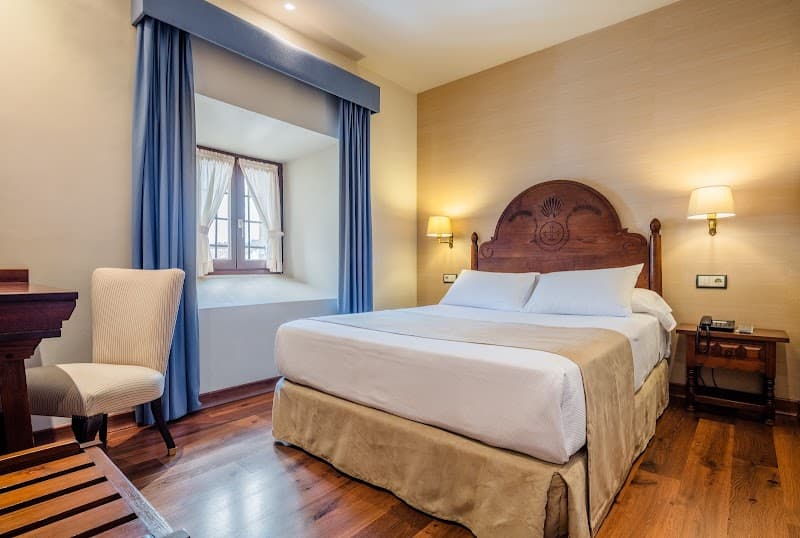 A cozy hotel room in Asturias, Spain, features warm wooden floors, a large bed with a carved headboard, soft lighting, and a serene window view, ideal for travelers seeking relaxation and comfort.