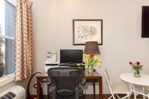 A cozy hotel room workspace features a modern office chair, desk with a TV and flowers, a small dining table, and warm curtains, ideal for travelers seeking comfort and productivity.