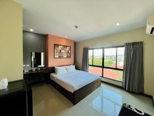 A cozy hotel room with a neatly made bed, vibrant orange accent wall, large windows offering a scenic view of rooftops and greenery, and modern furnishings creates an inviting atmosphere ideal for travelers seeking comfort and relaxation.