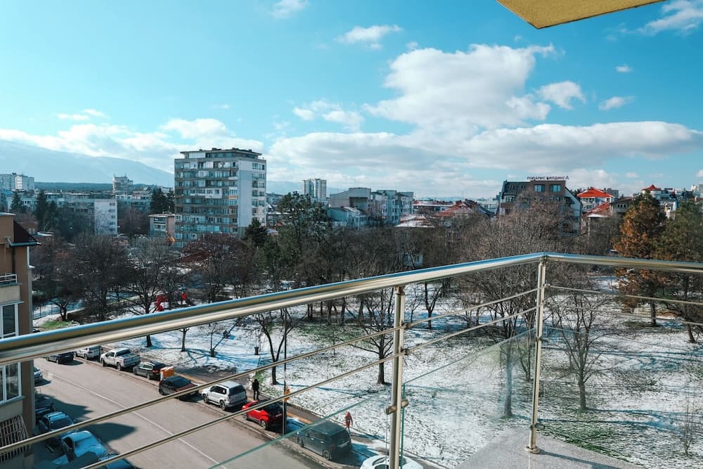 A serene winter cityscape viewed from a balcony railing, featuring snow-dusted trees, parked cars along a quiet street, and modern apartment buildings under a bright blue sky, ideal for urban winter retreats.
