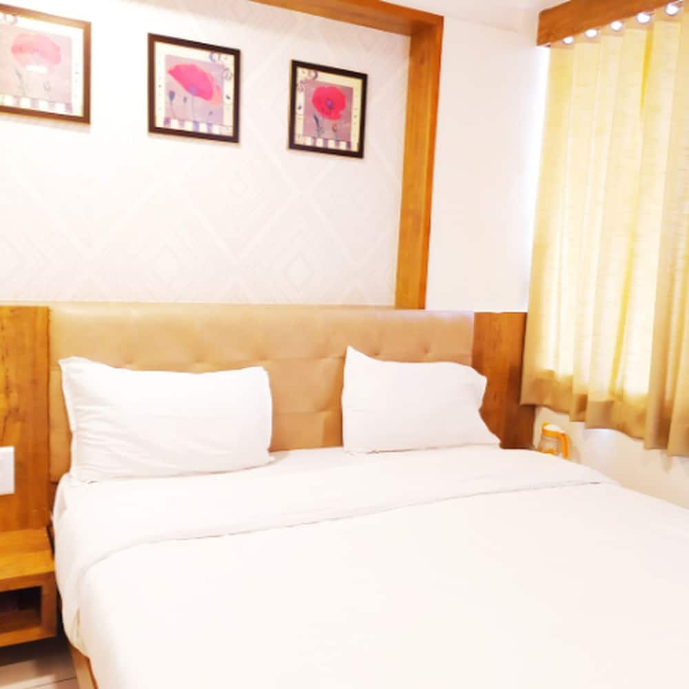A cozy hotel room features a neatly made bed with white linens, wooden accents, floral artwork on the walls, and soft yellow curtains, ideal for travelers seeking comfort and relaxation.