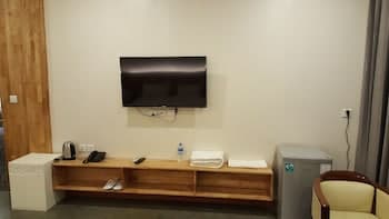 A cozy hotel room corner features a mounted TV above a wooden shelf holding towels, a remote, and water bottles, creating a relaxed atmosphere ideal for travelers seeking comfort and convenience.