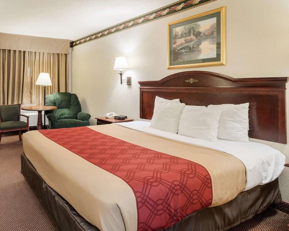 A cozy hotel room features a neatly made bed with red and beige bedding, a wooden headboard, comfortable seating by the window, and warm lighting, ideal for travelers seeking relaxation and comfort.