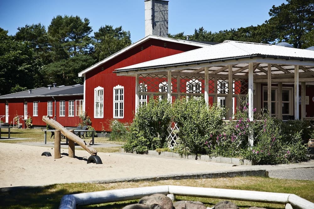 A charming red building with white trim and a covered porch stands amidst lush greenery, featuring a playful seesaw in the sandy foreground, ideal for family-friendly vacations and serene retreats.