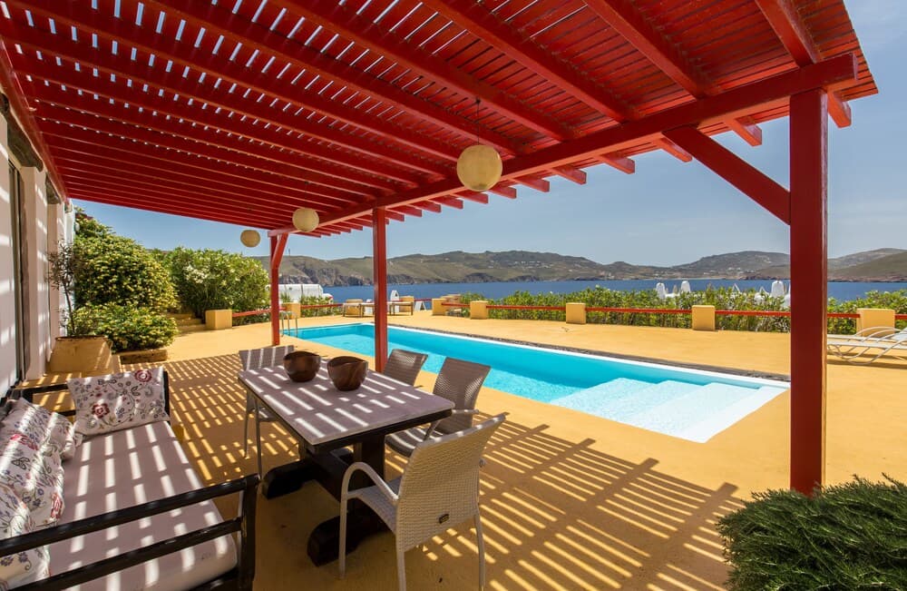 A serene coastal villa patio features a vibrant red pergola, comfortable seating, and a sparkling pool overlooking the tranquil sea, ideal for a luxurious vacation retreat.