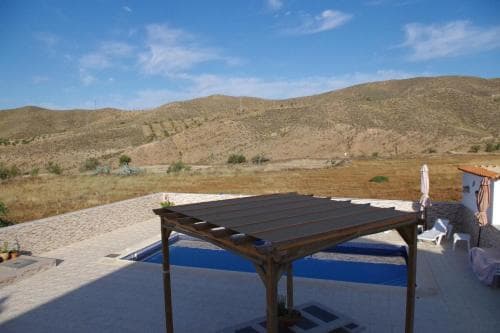 A serene outdoor patio with a wooden pergola overlooks a tranquil pool and expansive arid hills under a clear blue sky, ideal for a peaceful vacation retreat.