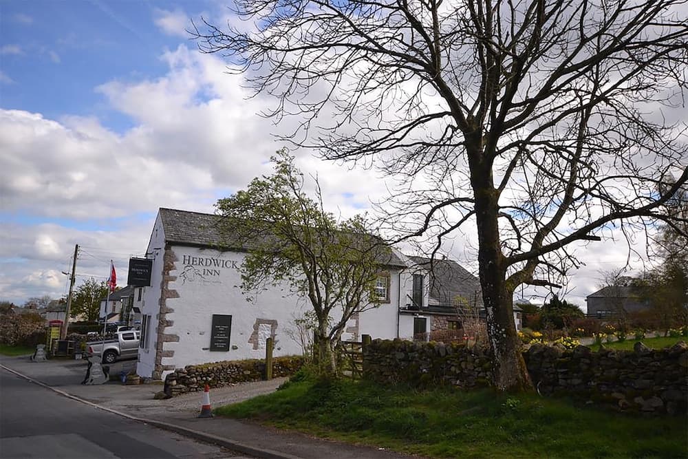 A quaint countryside pub named "Herdwick Inn" stands invitingly beside a stone wall under a partly cloudy sky, perfect for travelers seeking rustic charm and local hospitality.
