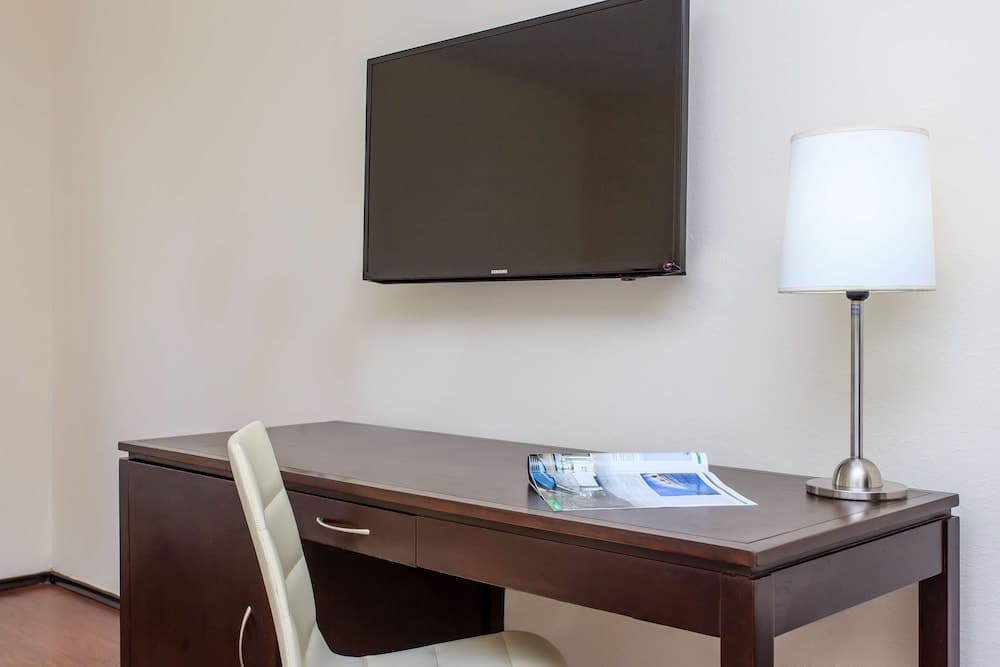 A cozy hotel room workspace features a sleek wooden desk with a white chair, a modern lamp, magazines, and a wall-mounted TV, ideal for travelers seeking comfort and productivity.