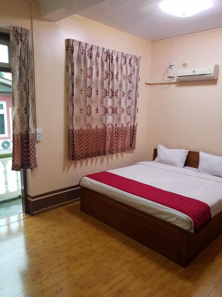 A cozy hotel room with a neatly made bed adorned with a red runner, patterned curtains, wooden flooring, and an air conditioner, ideal for travelers seeking comfort and simplicity.