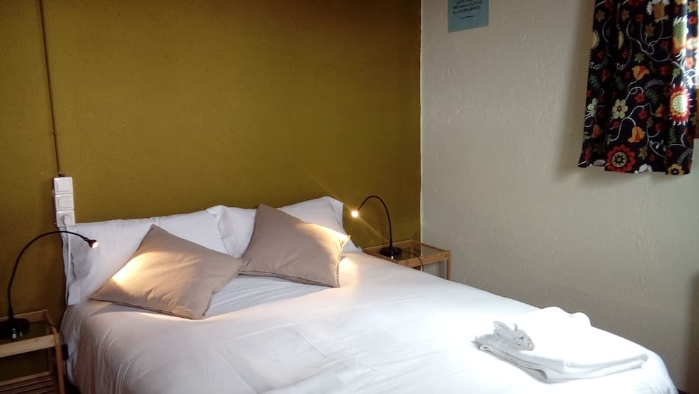A cozy hotel room features a neatly made bed with beige pillows against a mustard-yellow wall, flanked by wooden nightstands with lamps, creating a warm and inviting atmosphere ideal for travelers seeking comfort.