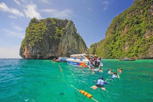Travelers enjoy snorkeling in vibrant turquoise waters surrounded by lush, towering cliffs under a sunny sky, ideal for adventure seekers exploring tropical destinations.