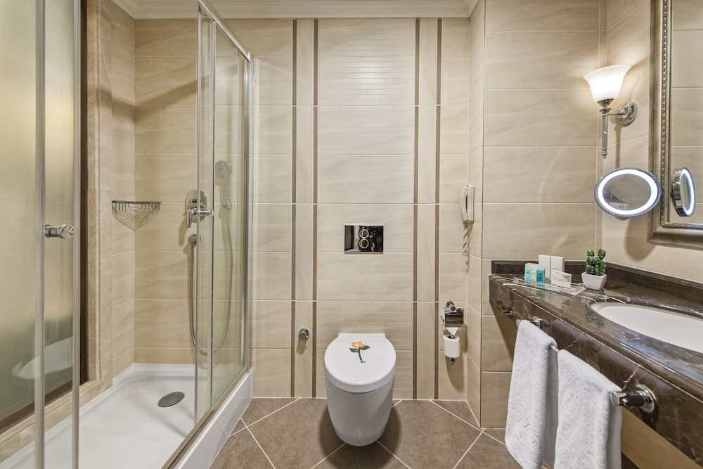 A serene hotel bathroom features beige tiled walls, a glass-enclosed shower, a modern toilet with a floral design, a sleek vanity with a sink and mirror, and soft lighting, ideal for travelers seeking comfort and cleanliness.