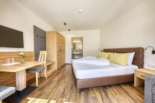 A cozy hotel room with warm wooden flooring, a neatly made bed with yellow pillows, a dining area, and a modern bathroom in the background, ideal for travelers seeking comfort and functionality.