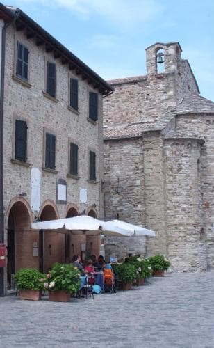 A serene European piazza features historic stone buildings, a quaint café with white umbrellas, and lush potted plants, ideal for travelers seeking a peaceful retreat in a charming medieval town.