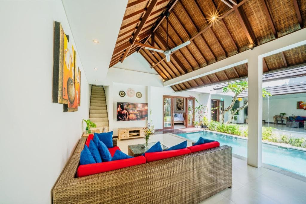 A cozy villa living area with vibrant red and blue cushions on a wicker sofa, a thatched roof ceiling, and an inviting indoor-outdoor pool view, perfect for a luxurious tropical getaway.
