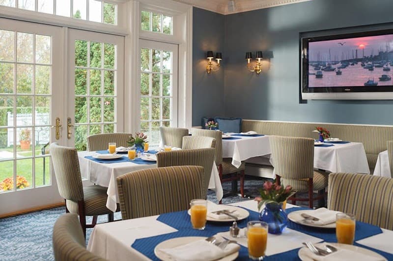 A cozy dining room at the Harbor Light Inn features elegant striped chairs, white tablecloths with blue accents, and large windows overlooking a serene garden, creating an inviting atmosphere perfect for travelers seeking a relaxing meal.