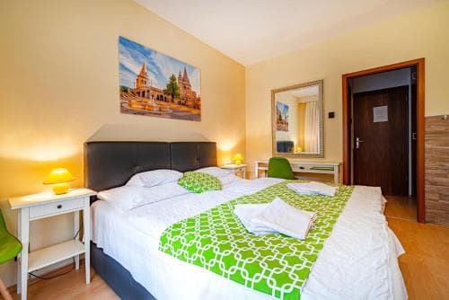 A cozy hotel room features a neatly made bed with green accents, warm lighting, and artwork of a cathedral, creating an inviting atmosphere ideal for travelers seeking comfort and relaxation.