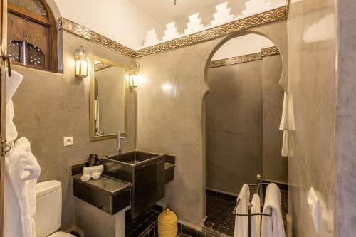 A cozy, rustic bathroom with arched doorways, decorative wall lights, and a stone sink exudes a serene, traditional ambiance ideal for travelers seeking cultural immersion in heritage accommodations.