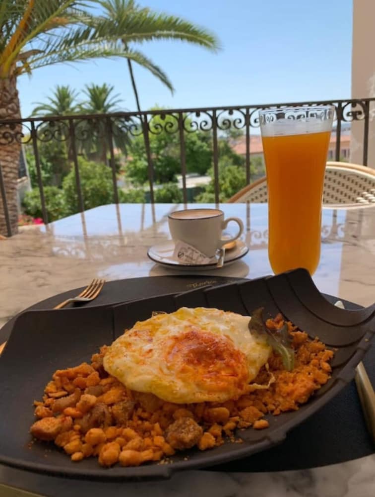 Enjoy a vibrant breakfast with a sunny Mediterranean vibe on a balcony adorned with palm trees, featuring a hearty dish topped with an egg, a refreshing orange juice, and a cup of coffee, perfect for a leisurely morning getaway.
