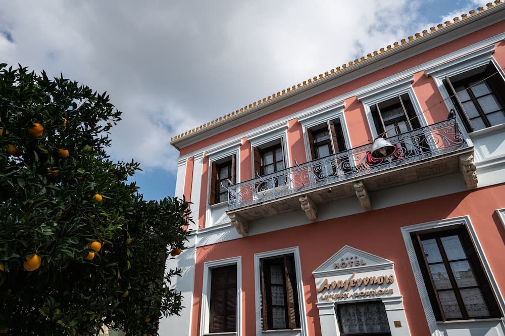 A charming boutique hotel with a pink facade and ornate balcony overlooks a vibrant orange tree under a partly cloudy sky, ideal for travelers seeking a picturesque European retreat.