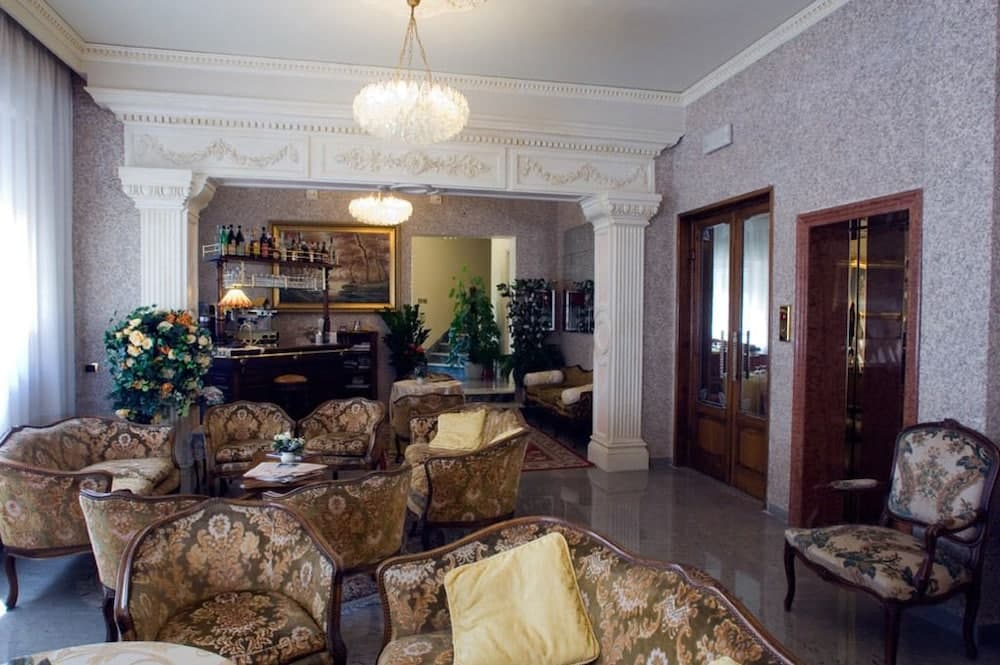 An elegant hotel lobby features ornate floral-patterned armchairs, a chandelier, decorative columns, and lush greenery, creating a luxurious and inviting atmosphere ideal for travelers seeking comfort and sophistication.