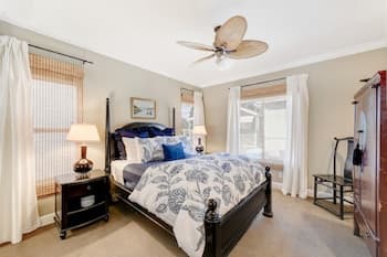 A cozy bedroom with a neatly made bed adorned in blue floral bedding, flanked by nightstands with lamps, features soft natural light filtering through sheer curtains, creating a serene atmosphere ideal for travelers seeking relaxation.