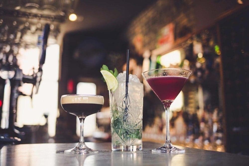 A cozy bar scene featuring three vibrant cocktails on a sleek countertop, set against a softly lit backdrop of shelves stocked with bottles, perfect for travelers seeking a relaxing nightlife experience.