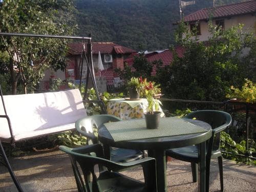 A cozy outdoor patio with a green table set for relaxation, surrounded by lush greenery and rustic houses, evokes a serene vacation vibe perfect for travelers seeking tranquility in a countryside retreat.