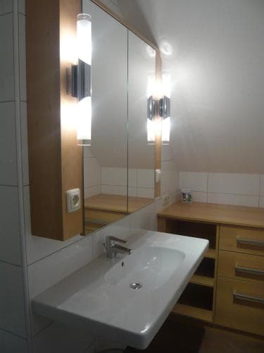 A modern bathroom features a sleek white sink, wooden cabinets, and mirrored storage illuminated by bright wall lights, creating a clean and functional space ideal for travelers seeking comfort in accommodations.