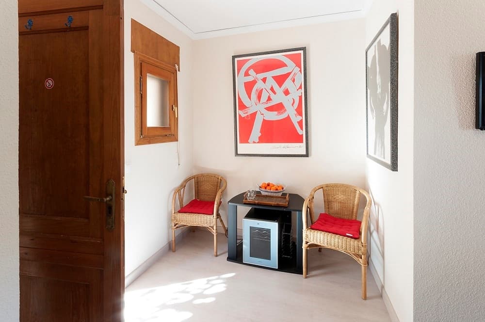 A cozy indoor lounge area features wicker chairs with red cushions, a small table with fruit and books, vibrant wall art, and natural light streaming in, ideal for travelers seeking a relaxing retreat space.